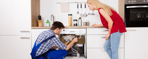 A man in blue overalls repairs a dishwasher in a modern kitchen while a woman in a red top and jeans watches attentively, considering advice from product liability attorneys. Various kitchen utensils and appliances are visible on the counter behind them; Baltimore County Product Liability Attorney concept image