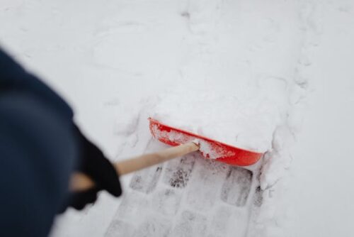 A person is removing snow with a red snow shovel, clearing a path on a snowy surface while wearing dark gloves and outerwear.