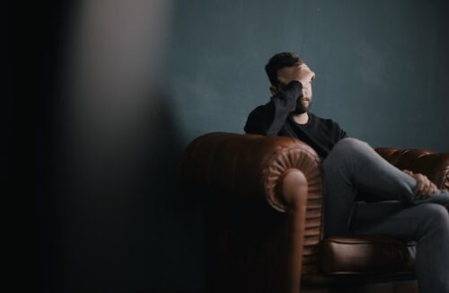 A person sits on a brown leather couch with one hand covering part of their face, appearing stressed or deep in thought, possibly struggling with Post Traumatic Stress Disorder, against a dark green wall.