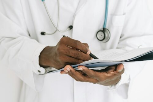 A doctor in a white coat and stethoscope writes notes on a clipboard, focusing on the hands and torso, as they update medical records against a plain background.