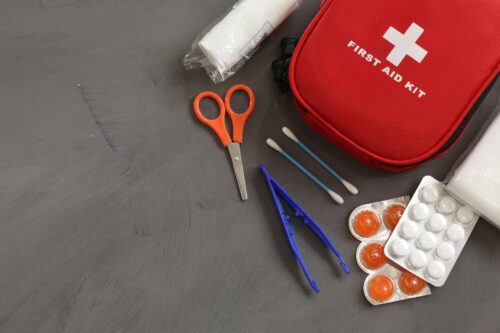 A red first aid kit with a white cross, scissors, tweezers, cotton swabs, bandages, and blister packs of pills laid out on a gray surface—ideal for treating minor wounds and burn injuries.