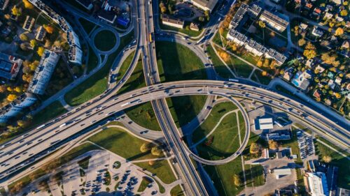 Aerial view of a large multi-lane highway interchange with overpasses, green spaces, and surrounding buildings in an urban area. Cars are visible on the roads while sunlight casts shadows—no sign of any road rage accident.