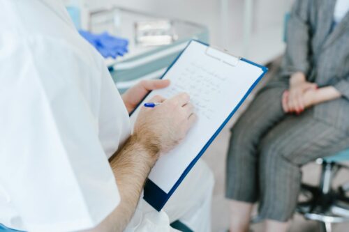 A person in a white coat writes on a clipboard with a pen, while another in a gray suit sits nearby with hands folded, suggesting a professional consultation, possibly discussing topics such as medical liens.