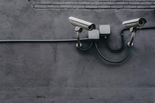 Two security cameras, ready to capture footage, are mounted side by side on a gray concrete wall. Black cables and metal junction boxes connect them, with a section of exposed brick visible near the top.