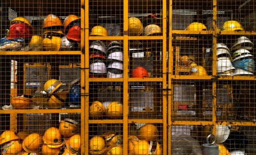 Rows of yellow metal shelves filled with various construction helmets, mostly yellow, with a few orange, white, and red ones. The wire mesh covering the shelves helps prevent workplace accidents by keeping helmets secure and accessible.
