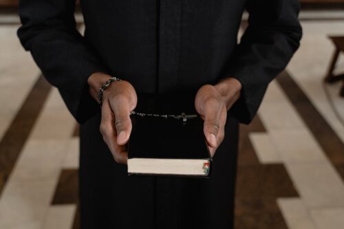 A person in a black robe, possibly a victim, holds a closed Bible with a rosary draped over it, standing indoors on a tiled floor—an image resonant with recent discussions about clergy abuse in Maryland.