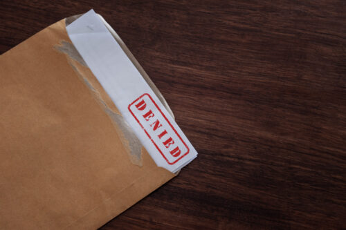 A torn brown envelope on a wooden surface reveals a white letter with a red "DENIED" stamp at the top, indicating a claim denied in Maryland.