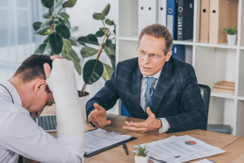 A man with a bandaged arm sits at a desk, looking distressed, as another man in a suit discusses his injury and return to work. Papers, a laptop, and office supplies are spread out in the modern office setting.