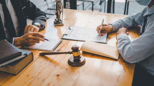 Two people sit at a wooden desk with legal documents, a gavel, and Lady Justice, discussing how their personal injury case could be settled without court as one writes on a clipboard and the other gestures with a pen.