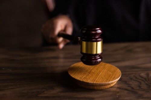 A hand holds a judge’s gavel above a wooden sound block on a dark wooden table, symbolizing justice or legal proceedings in a case involving allegations of abusing 11 boys.